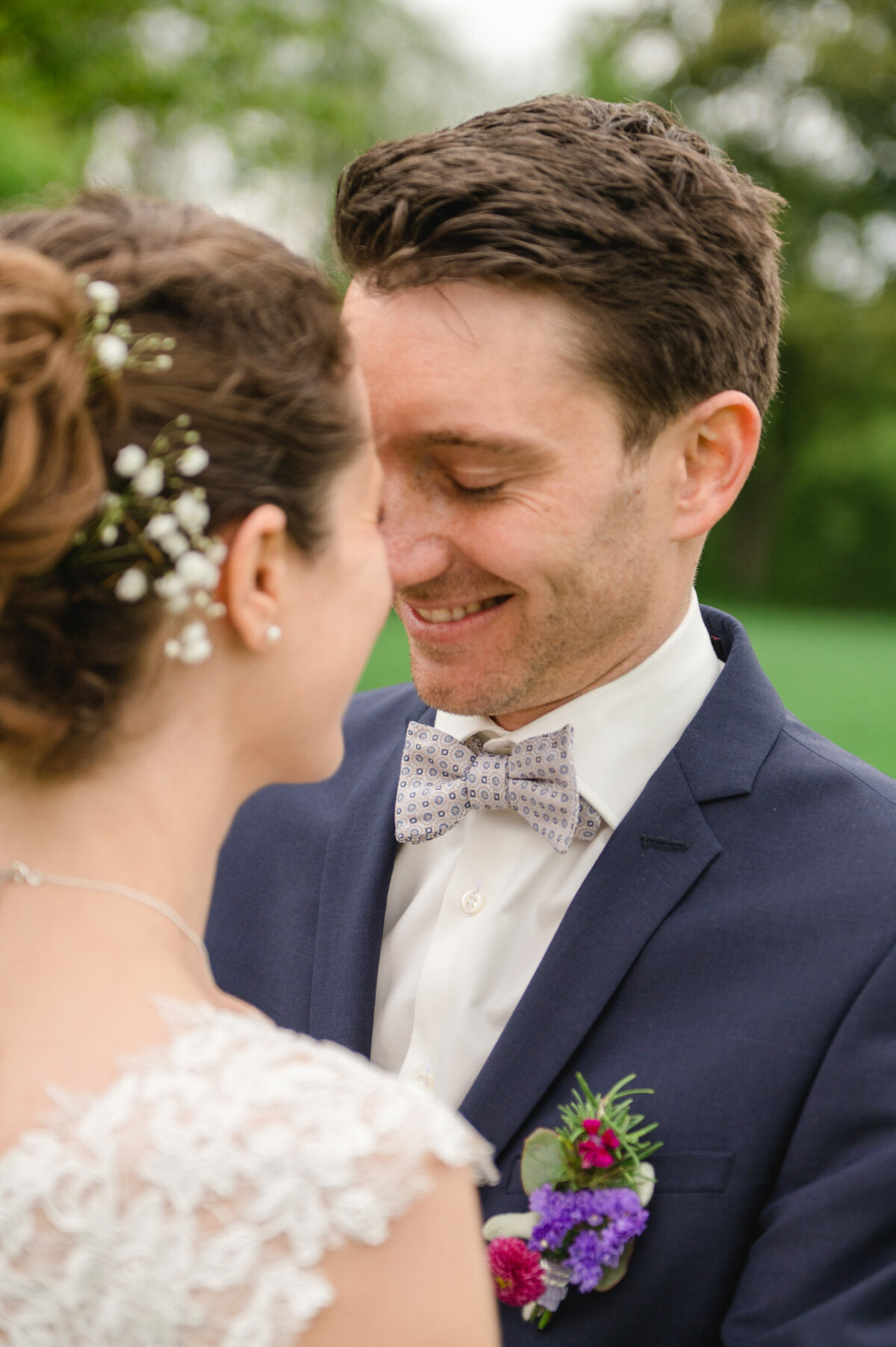 Eine Braut und ein Bräutigam lächeln sich im Freien an. Der Bräutigam trägt einen blauen Anzug und eine Blumenansteckblume und die Braut trägt floralen Haarschmuck.