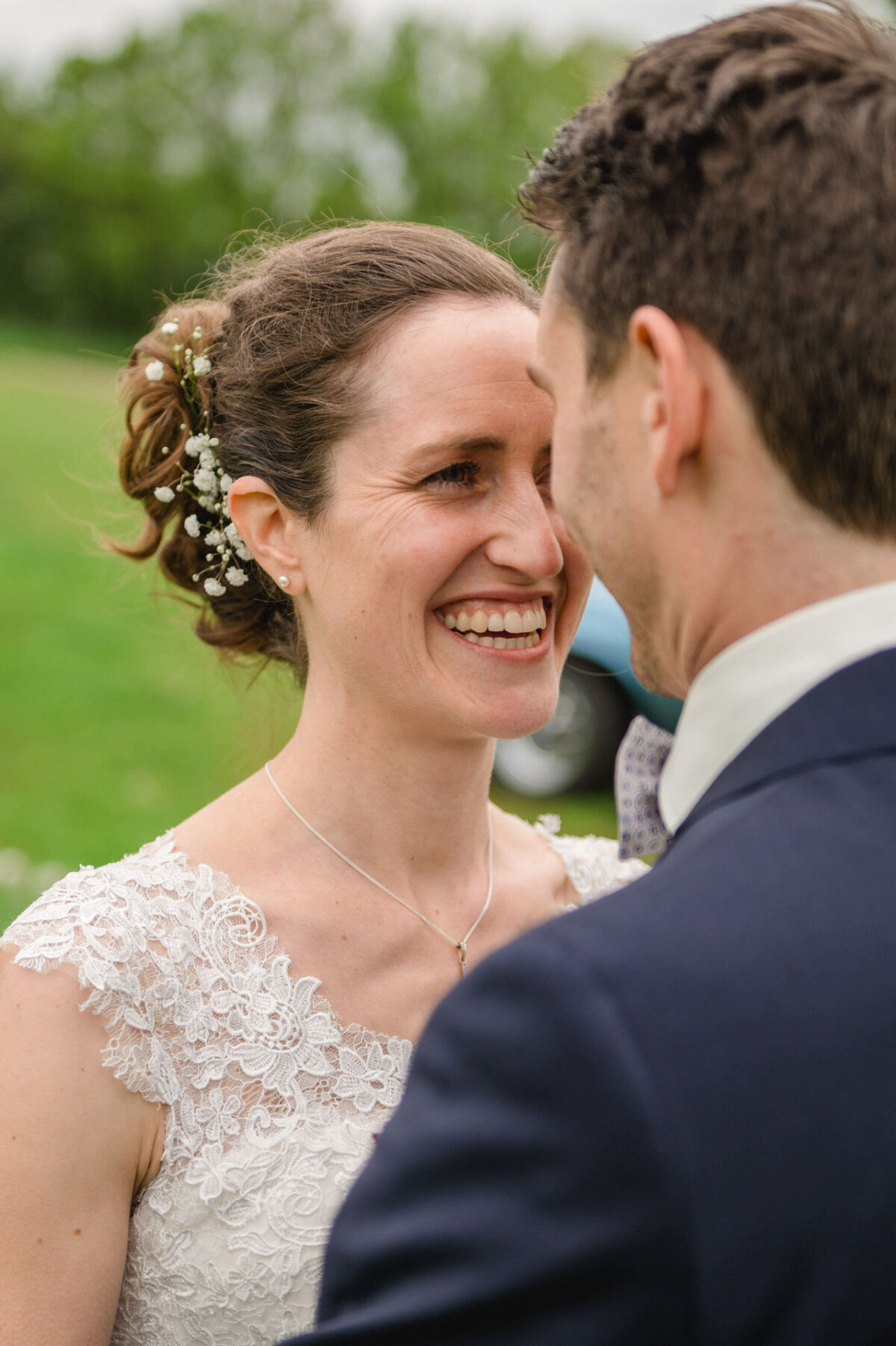 Eine lächelnde Frau in einem weißen Spitzenkleid steht im Freien einem Mann im Anzug gegenüber.