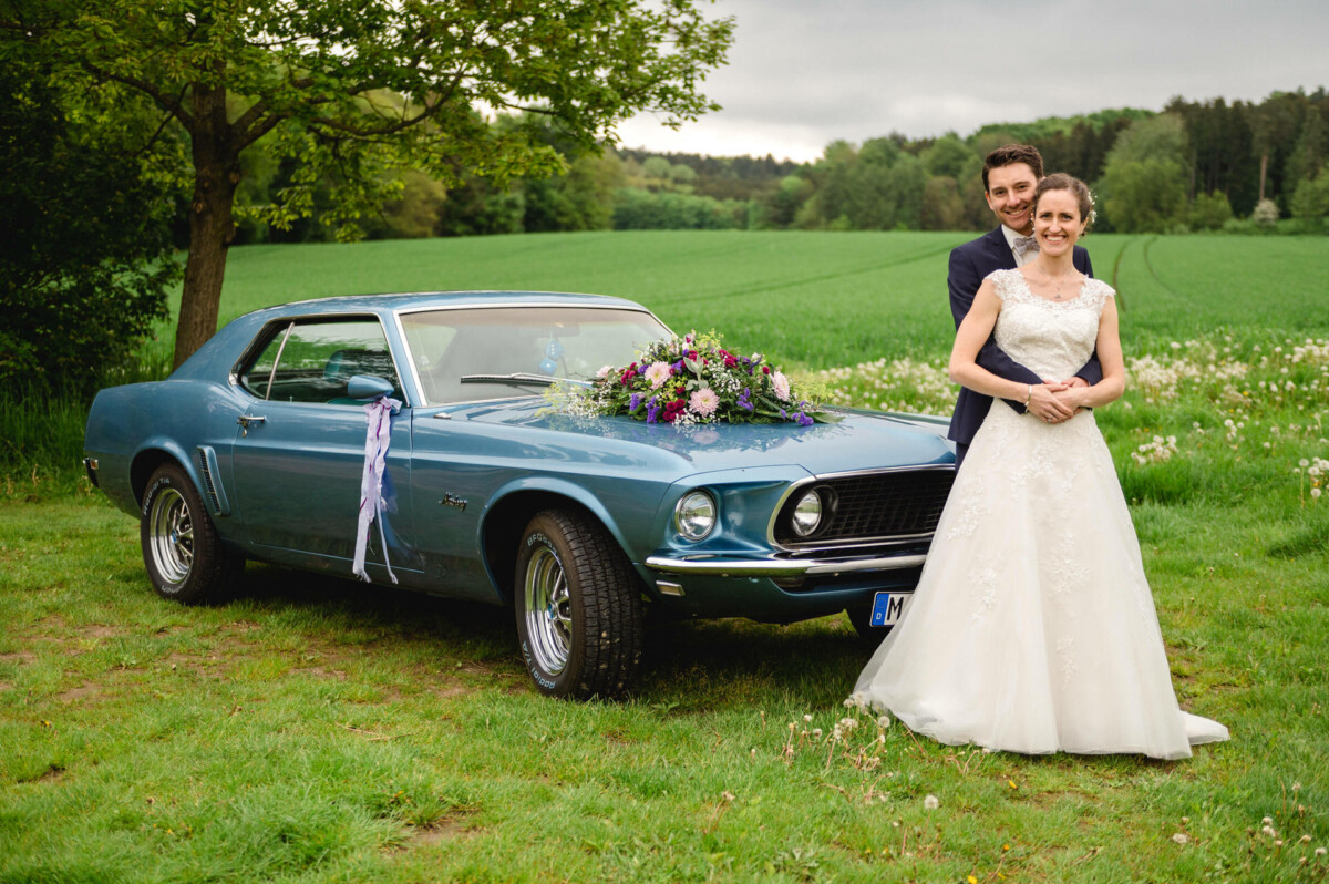 Braut und Bräutigam posieren vor einem mit Blumen geschmückten blauen Oldtimer auf einer Wiese mit Bäumen im Hintergrund.