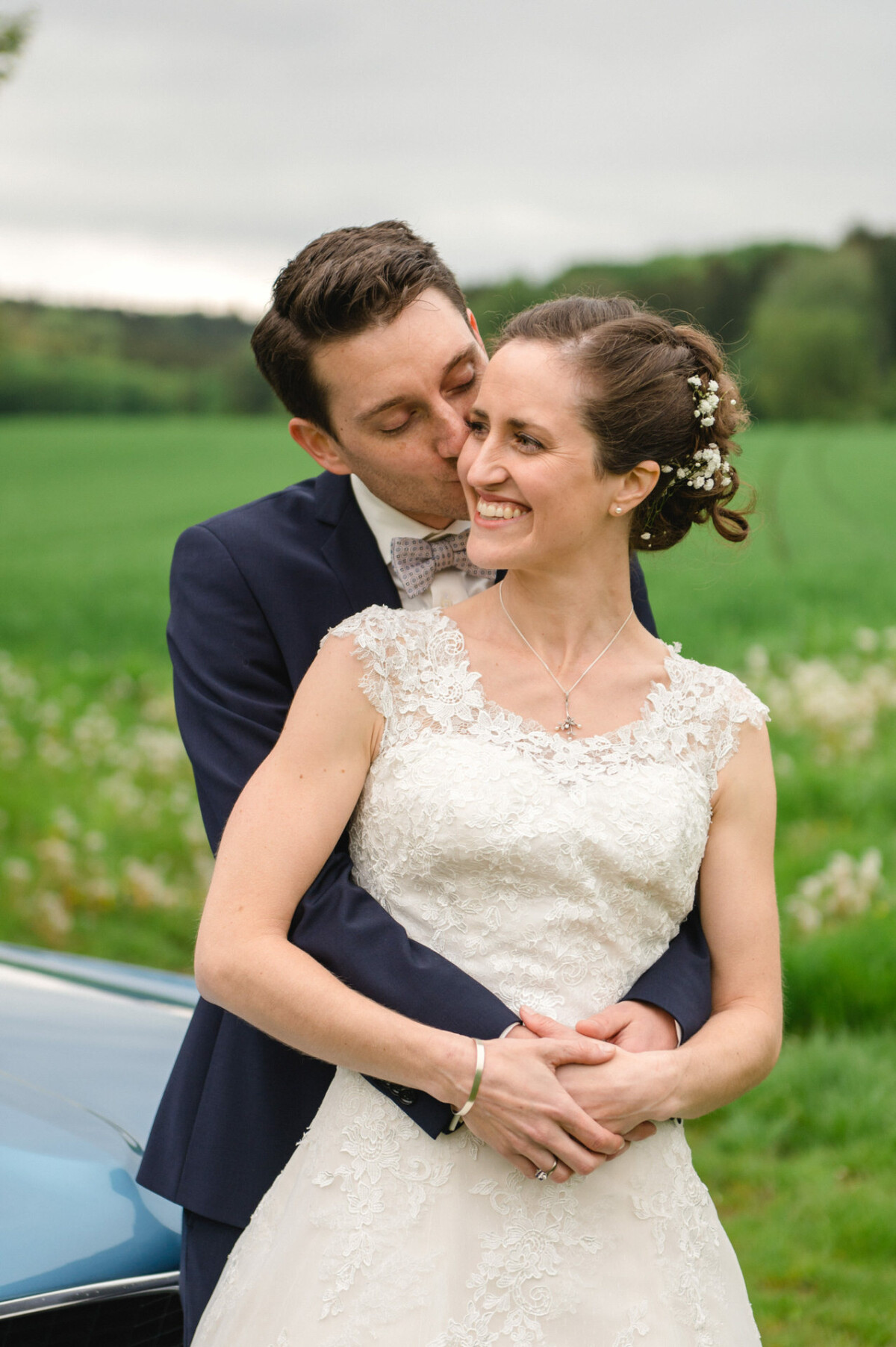 Ein Bräutigam im marineblauen Anzug küsst eine lächelnde Braut im Spitzenkleid. Sie stehen draußen auf einer grünen Wiese mit Bäumen im Hintergrund.