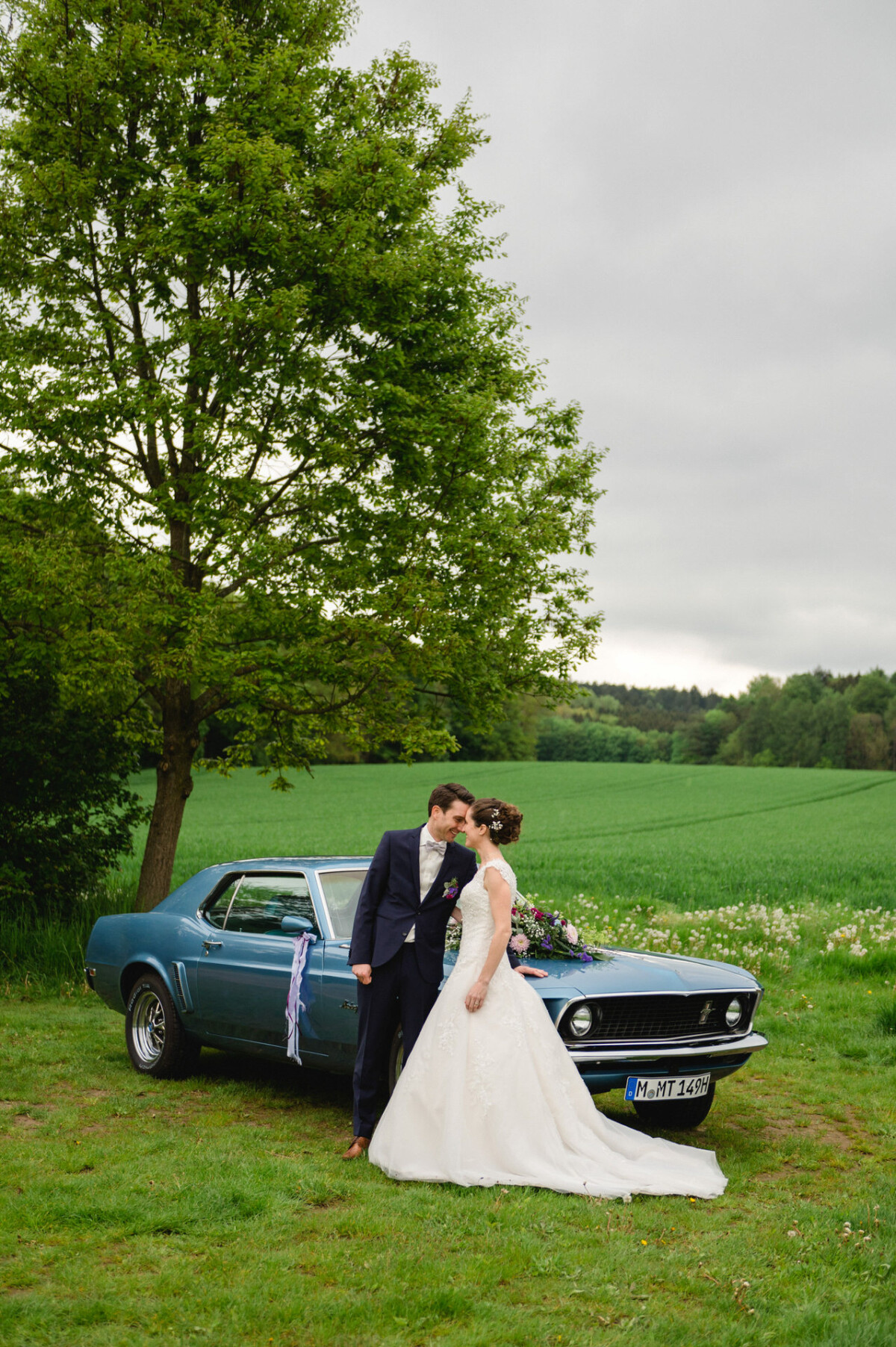Braut und Bräutigam stehen neben einem blauen Oldtimer auf einer Wiese mit Bäumen. Die Braut trägt ein weißes Kleid und der Bräutigam einen dunklen Anzug.