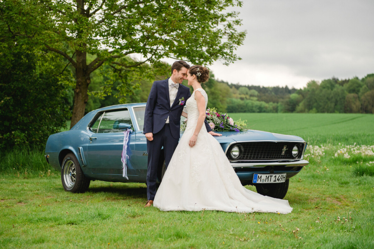 Ein Paar im Hochzeitskleid steht neben einem Oldtimer auf einer Wiese, im Hintergrund sind ein Baum und Grün zu sehen.
