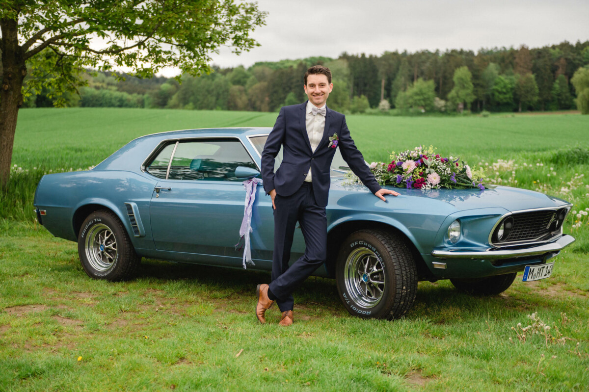 Ein Mann im Anzug lehnt an einem blauen Oldtimer mit einem Blumenarrangement auf der Motorhaube, der auf einer Wiese mit Bäumen im Hintergrund geparkt ist.