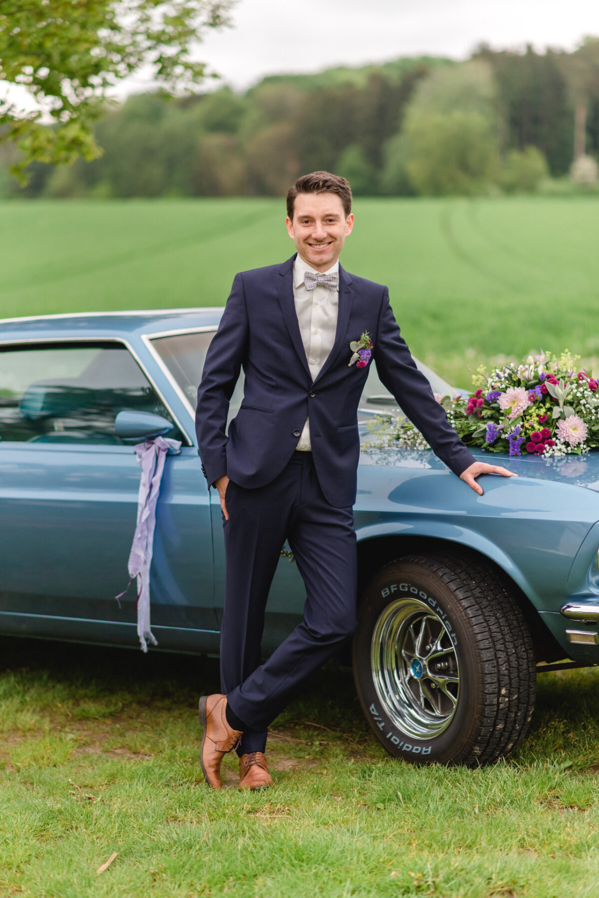 Ein Mann im Anzug steht lächelnd neben einem blauen, mit Blumen geschmückten Oldtimer auf einer Rasenfläche mit einem Feld im Hintergrund.