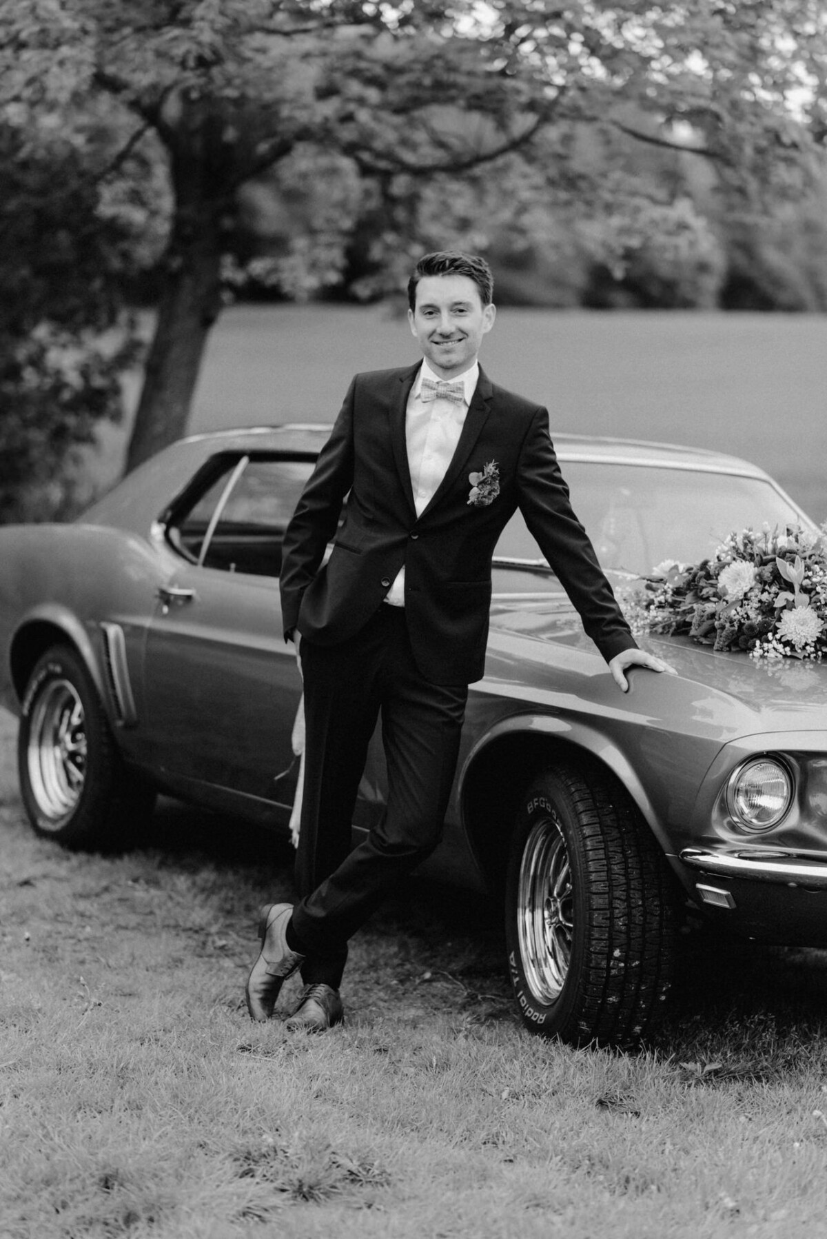 Junger Mann im Anzug lehnt im Freien an einem Oldtimer mit einem Blumenarrangement auf der Motorhaube.