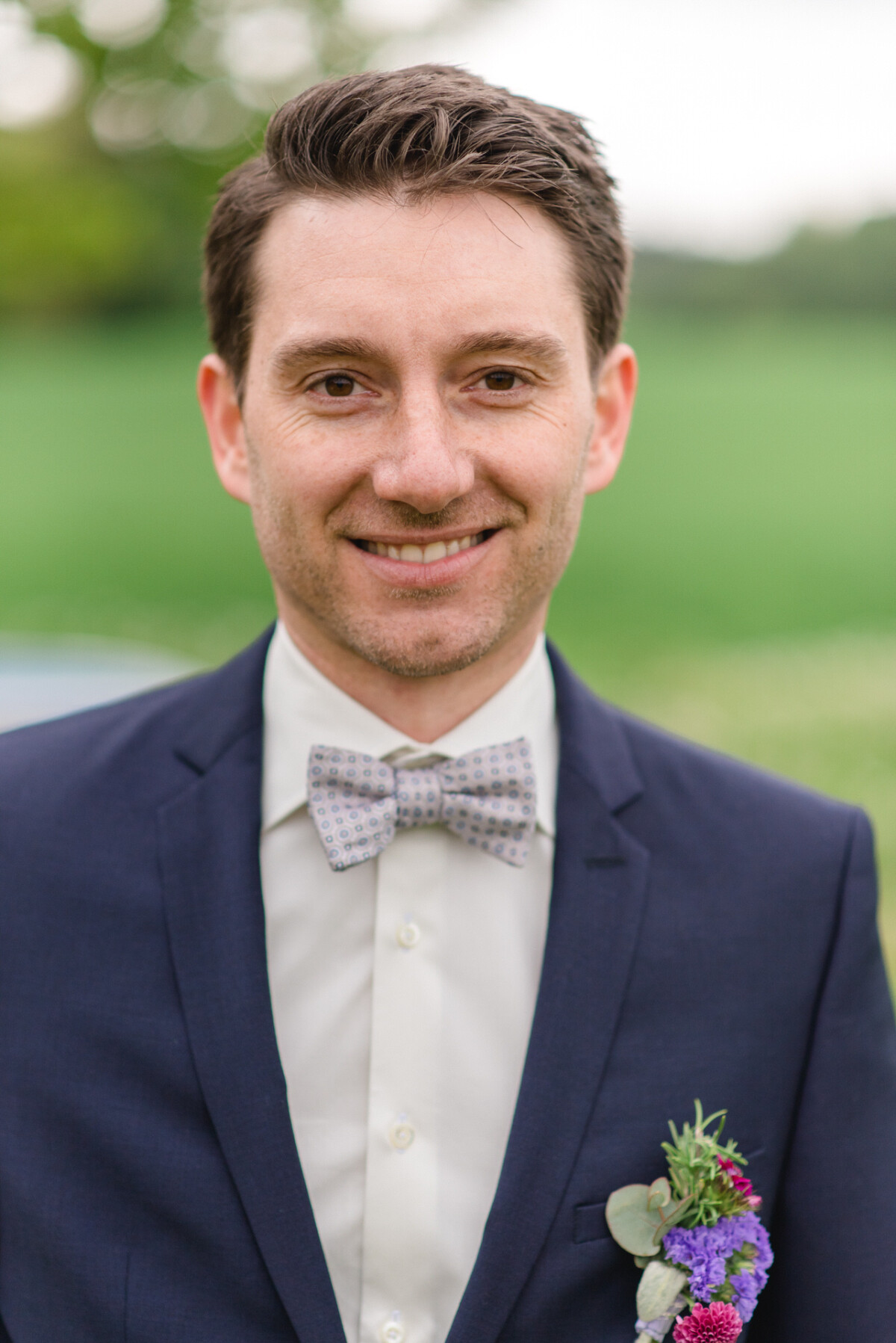 Ein Mann in einem marineblauen Anzug und einer gemusterten Fliege steht lächelnd im Freien. An seinem Revers trägt er eine Blumenboutonniere, der Hintergrund ist unscharf und grün.