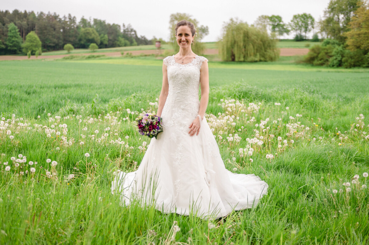 Eine Braut in einem Spitzenbrautkleid steht auf einer leuchtend grünen Wiese und hält einen Blumenstrauß in der Hand.