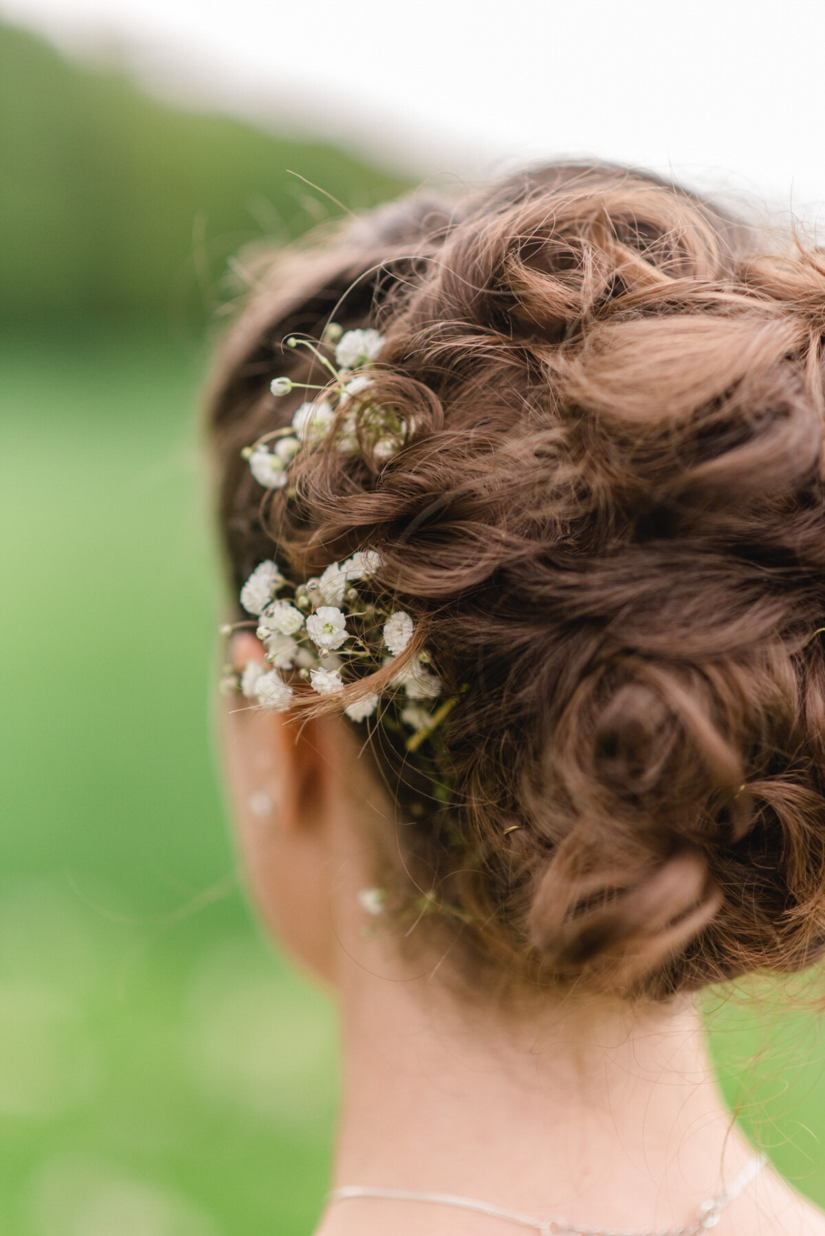 Nahaufnahme der braunen Haare einer Frau in einer Hochsteckfrisur mit kleinen weißen Blumen vor einem unscharfen grünen Hintergrund.