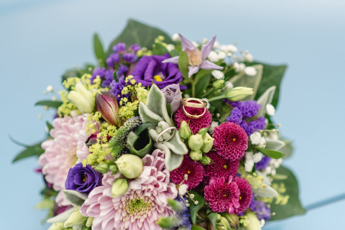 Ein lebendiger Blumenstrauß mit rosa und lila Blumen, Grünpflanzen und zwei Eheringen oben auf einem hellblauen Hintergrund.