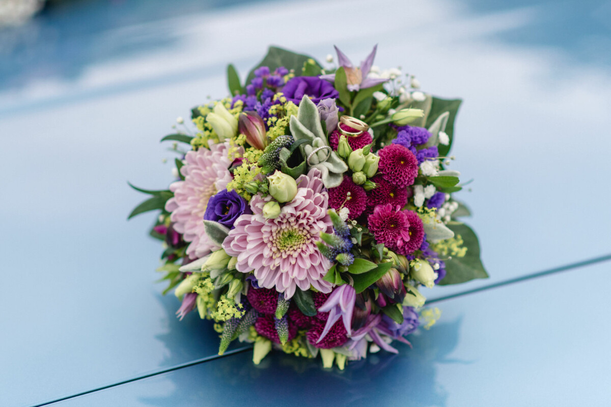 Ein farbenfroher Blumenstrauß mit violetten, rosa und weißen Blüten auf hellblauem Untergrund. Zwischen den Blüten ist ein goldener Ring platziert.