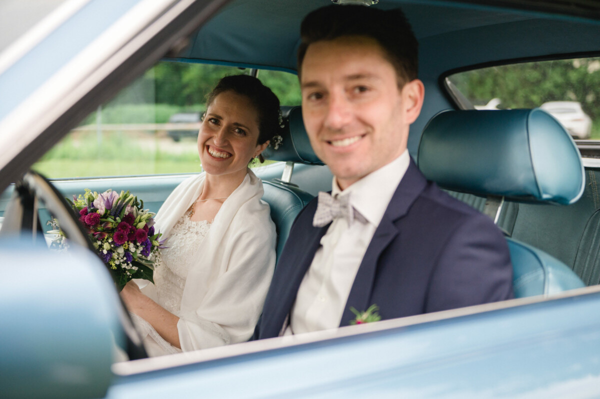 Braut und Bräutigam sitzen lächelnd im Auto. Die Braut hält einen Blumenstrauß in der Hand.