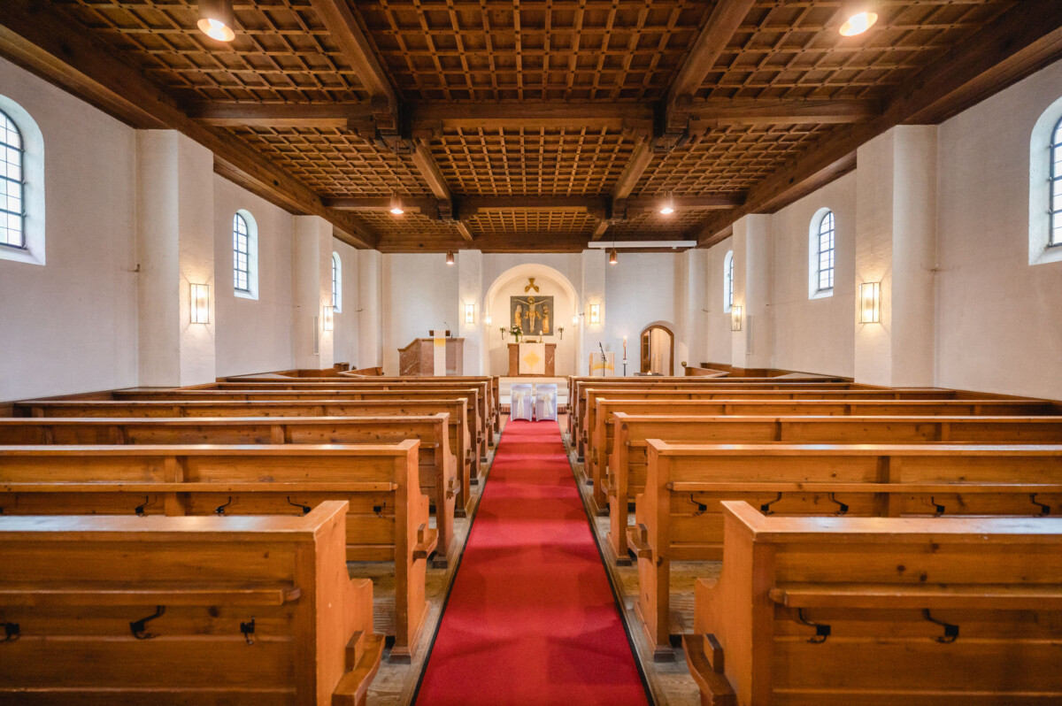 Innenansicht einer kleinen Kirche mit Holzbänken, einem mit rotem Teppich ausgelegten Mittelgang und einem Altar an der Vorderseite, umgeben von Bogenfenstern und einer hölzernen Kassettendecke.