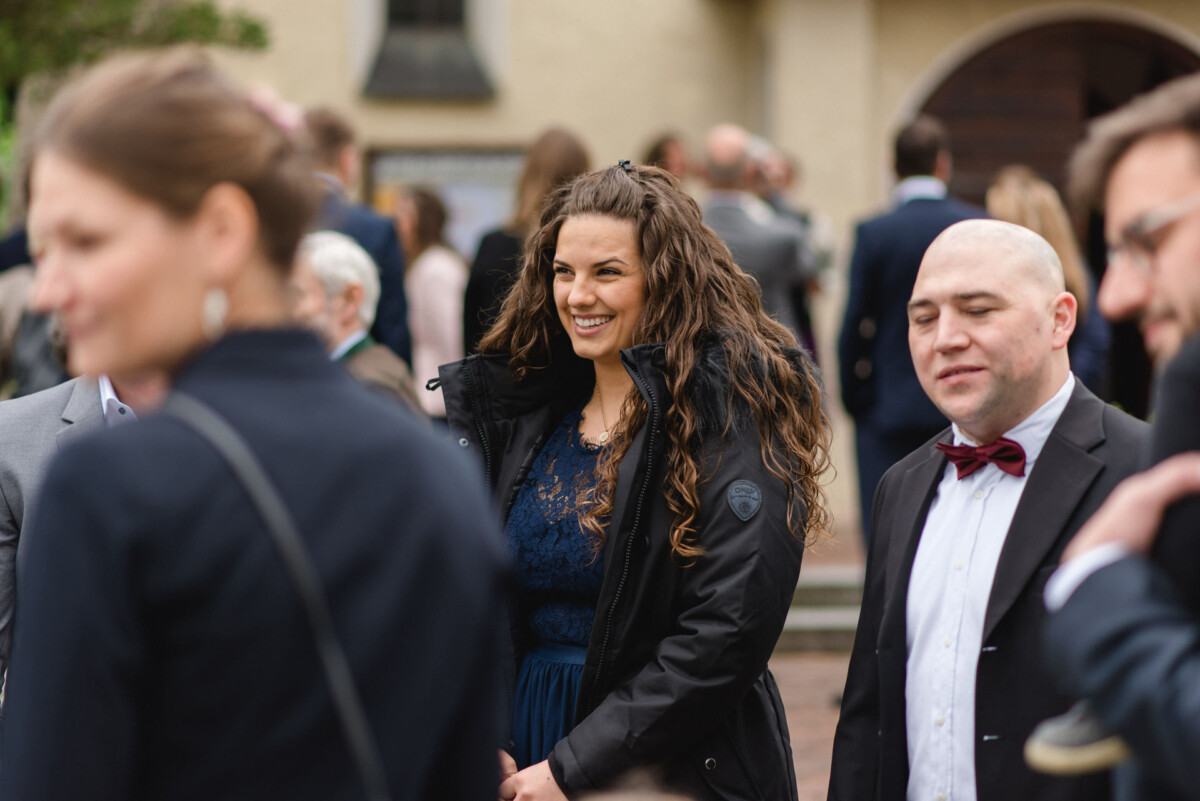 Eine Gruppe von Menschen in formeller Kleidung steht draußen bei einer Veranstaltung. Eine Frau in einem blauen Kleid und einem schwarzen Mantel lächelt.