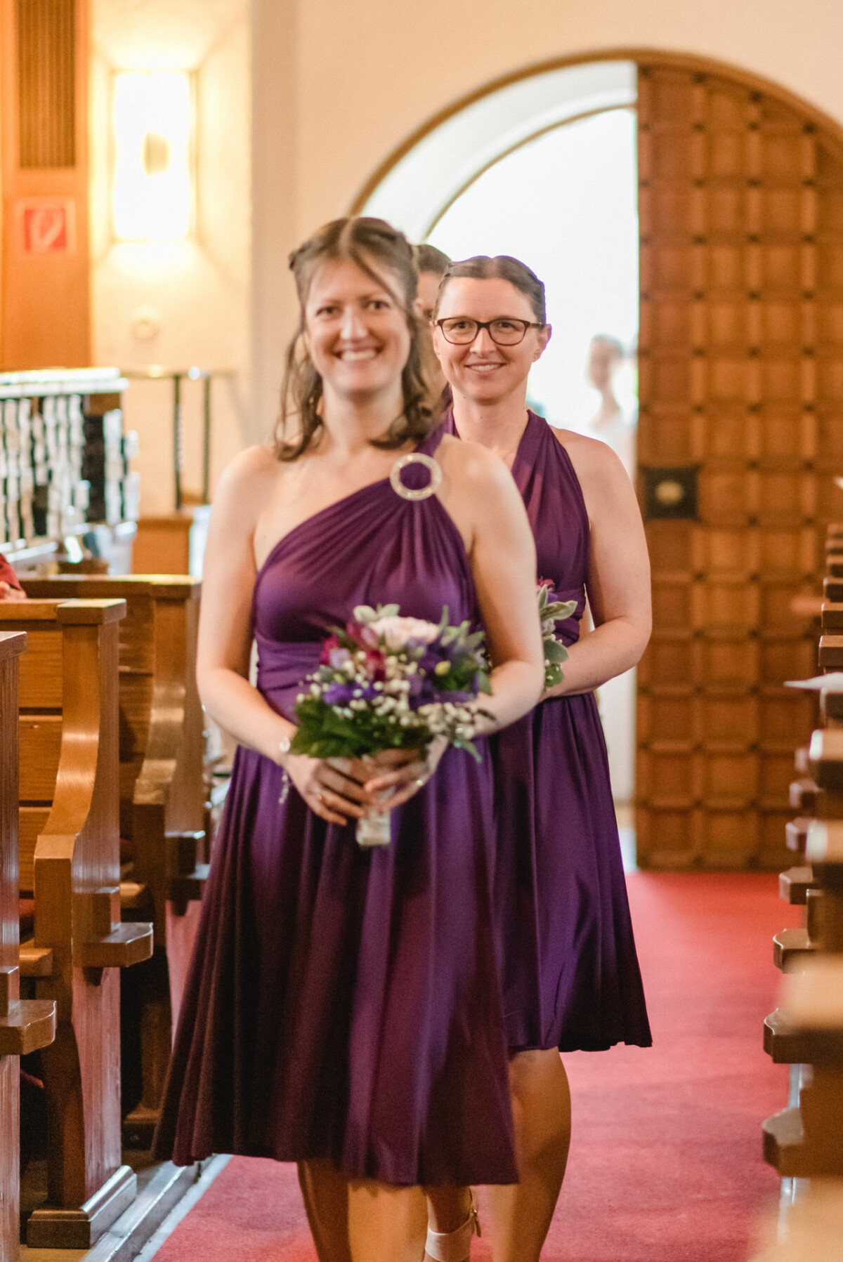 Zwei Frauen in lila Kleidern gehen in einer Kirche einen Gang entlang, jede hält einen Blumenstrauß in den Händen.