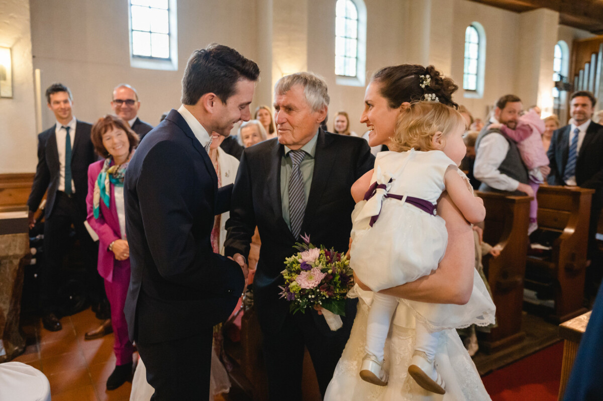 Eine Gruppe von Menschen in Abendgarderobe steht während einer Hochzeitszeremonie in einer Kirche. Eine Frau hält ein Kind, während sich zwei Männer in der Nähe die Hand schütteln.