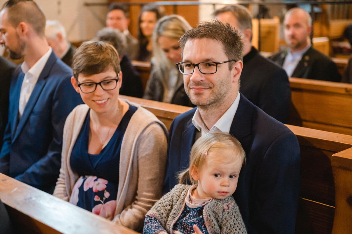 Ein Mann und eine Frau sitzen in einer Holzbank, der Mann hat ein Kleinkind auf dem Schoß, umgeben von anderen sitzenden Erwachsenen in einem Raum.