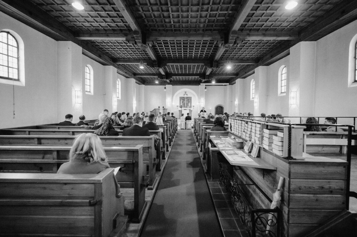 Ein Schwarzweißfoto von Menschen, die während eines Gottesdienstes in Holzbänken in einer Kirche sitzen und dem Altar zugewandt sind.