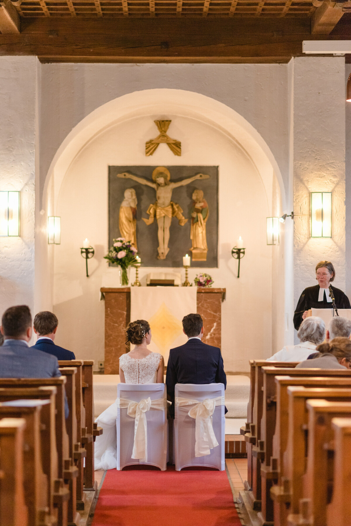 Ein Paar sitzt während einer kirchlichen Trauungszeremonie am Altar, mit Blick auf den Geistlichen und die Gäste, im Hintergrund sind ein Kruzifix und Blumenarrangements zu sehen.