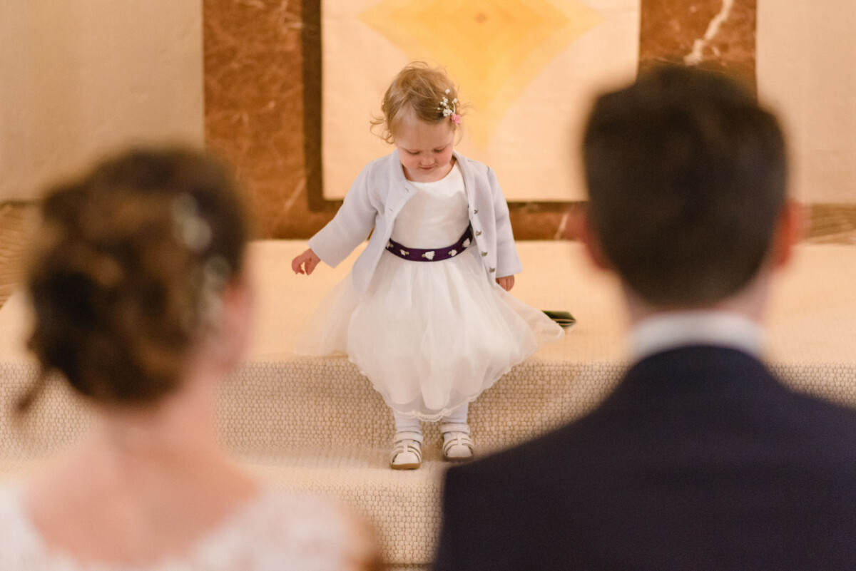 Ein kleines Kind in einem weißen Kleid steht auf einer Treppe und blickt dem Paar im Hochzeitskleid entgegen.