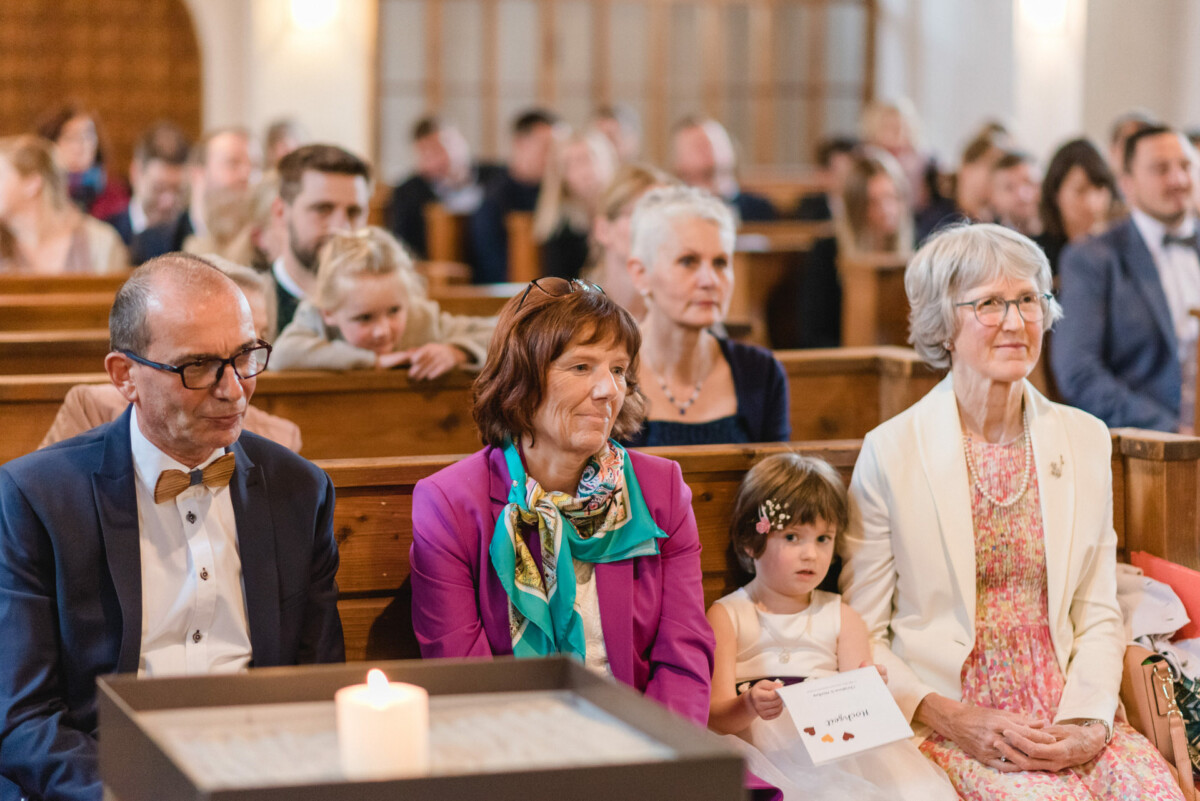 Menschen sitzen in Reihen von Holzbänken in einer Kirche, im Vordergrund brennt eine Kerze.