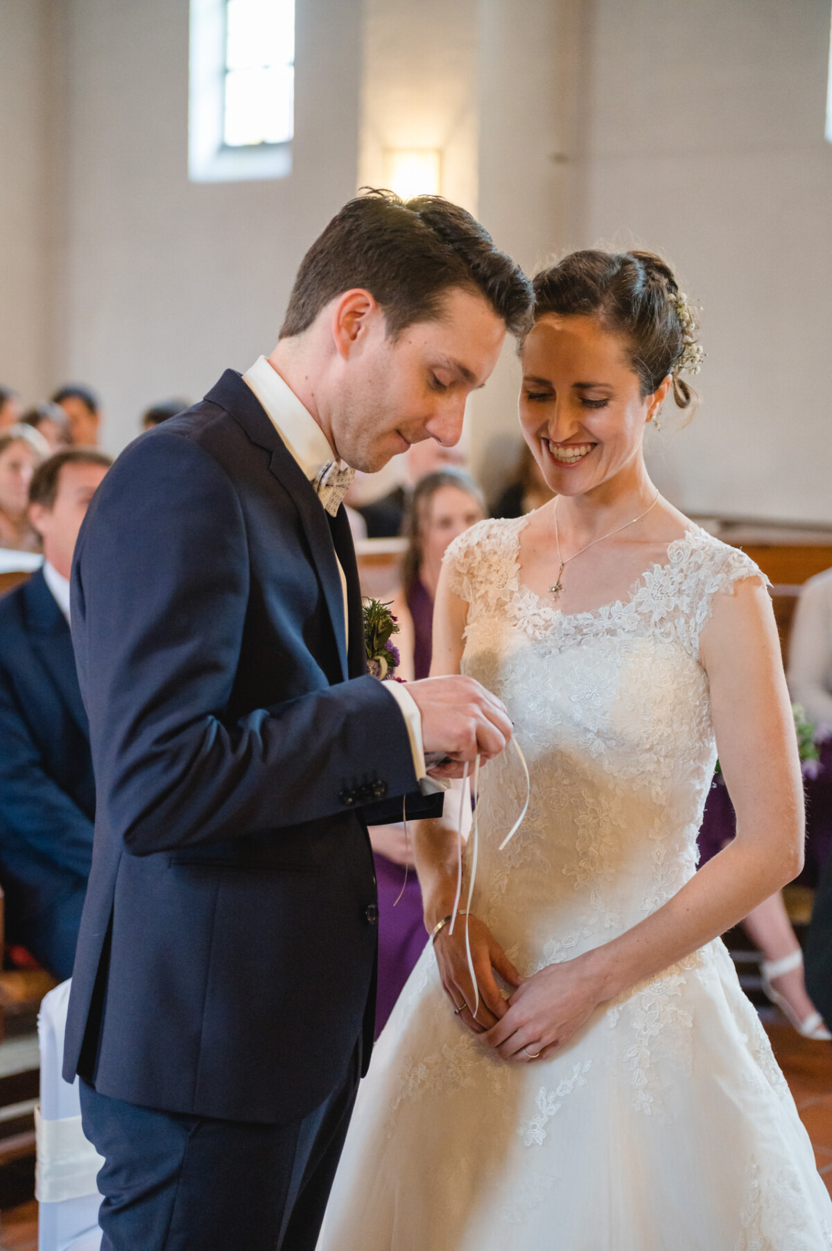 Braut und Bräutigam stehen zusammen in einer Kirche. Der Bräutigam hält ein Band und beide lächeln. Im Hintergrund sitzen Gäste.