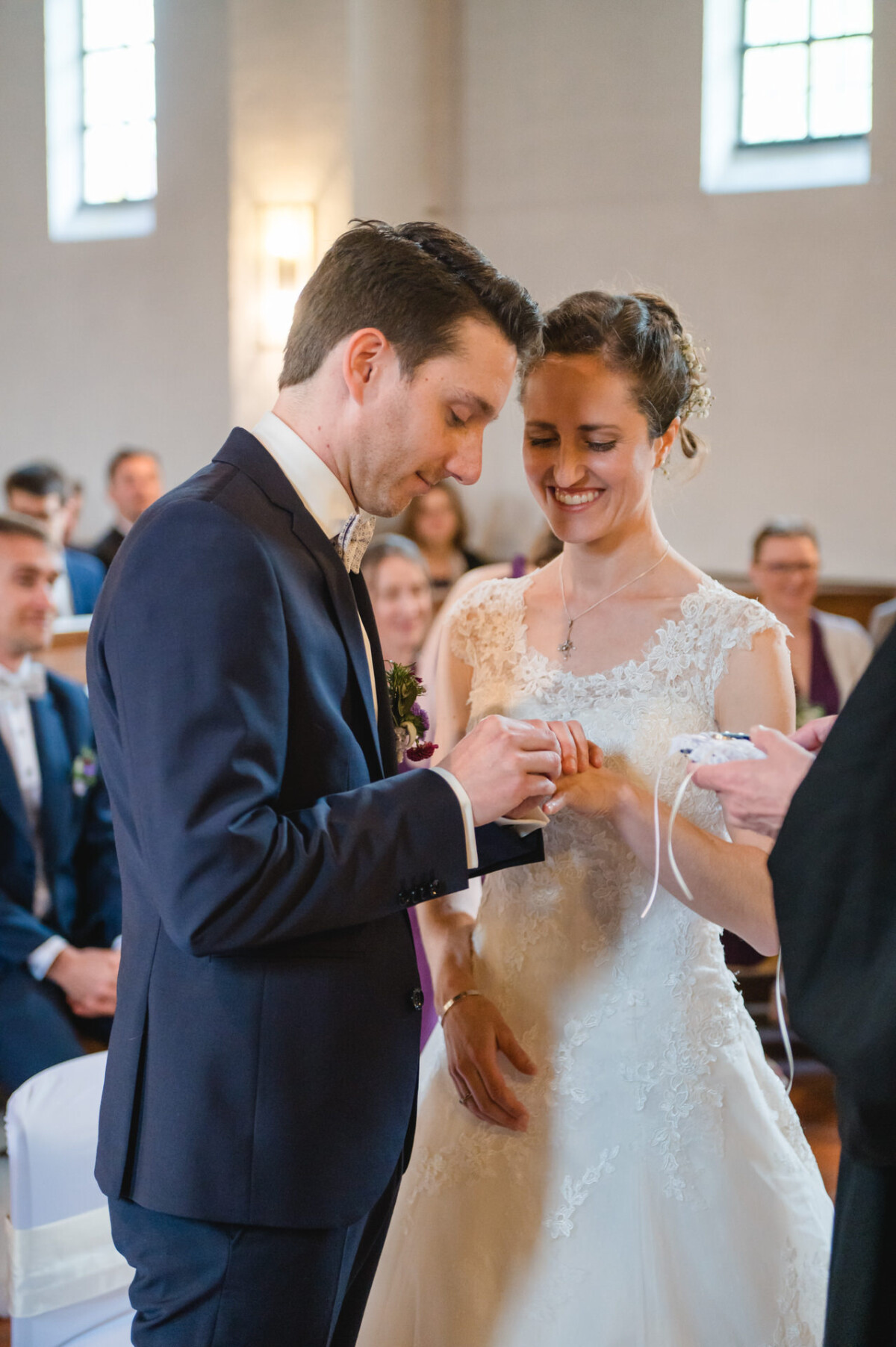 Ein Bräutigam steckt der Braut während einer Hochzeitszeremonie in einer Kirche, umgeben von sitzenden Gästen, einen Ring an den Finger.