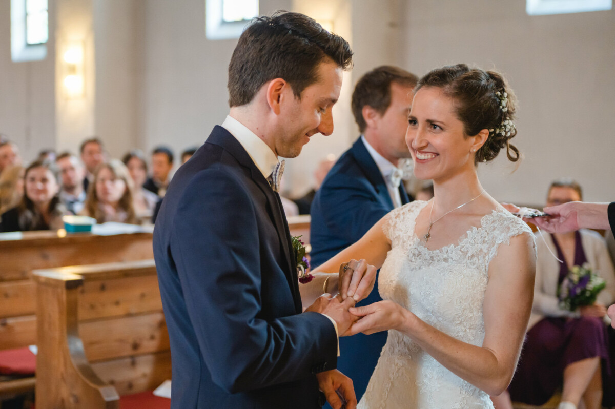 Ein Paar tauscht während seiner Hochzeitszeremonie in einer Kirche, umgeben von sitzenden Gästen, Ringe aus.