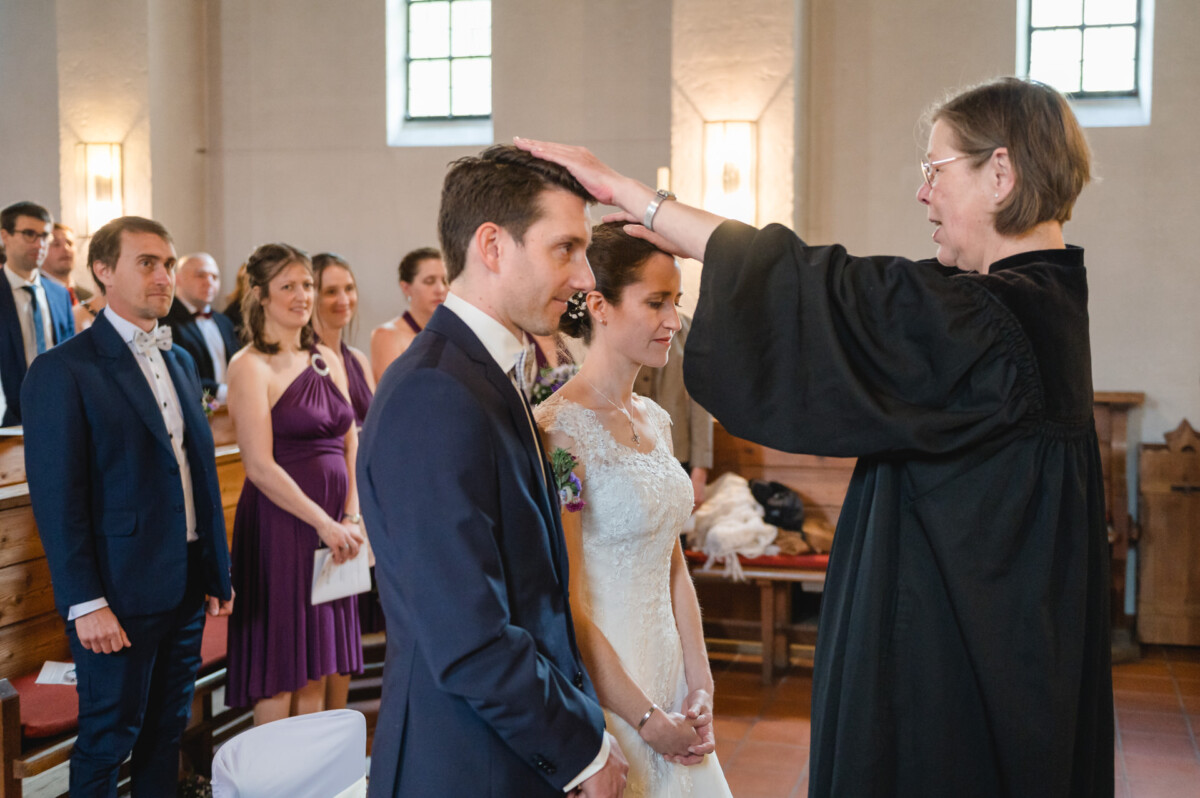 Ein Paar steht während einer kirchlichen Trauungszeremonie am Altar, während ein Pfarrer dem Bräutigam die Hand auf den Kopf legt. Im Hintergrund sitzen Gäste.