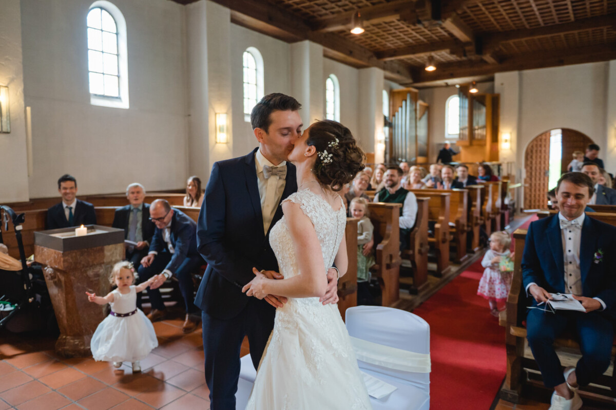 Ein Paar in Abendgarderobe küsst sich während der Hochzeitszeremonie in einer Kirche am Altar, während die Gäste in Holzbänken sitzen.