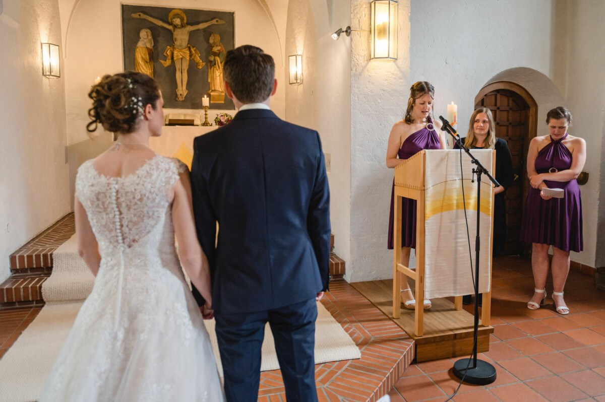 Ein Paar steht während einer Hochzeitszeremonie in einer Kirche zusammen, während eine Frau am Rednerpult vorliest. In der Nähe stehen zwei weitere Frauen in lila Kleidern.