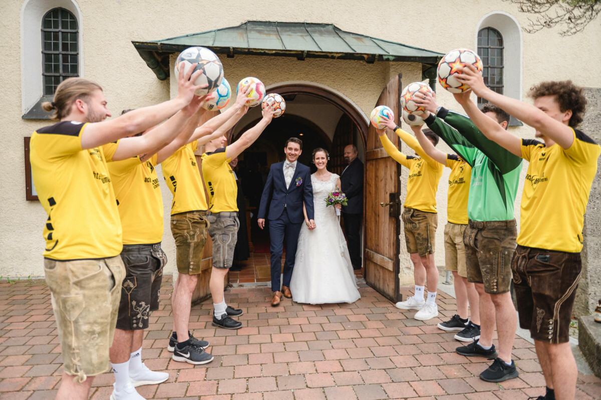 Braut und Bräutigam verlassen ein Gebäude durch einen Torbogen, der aus Männern in gelben Hemden besteht, die Fußbälle halten.