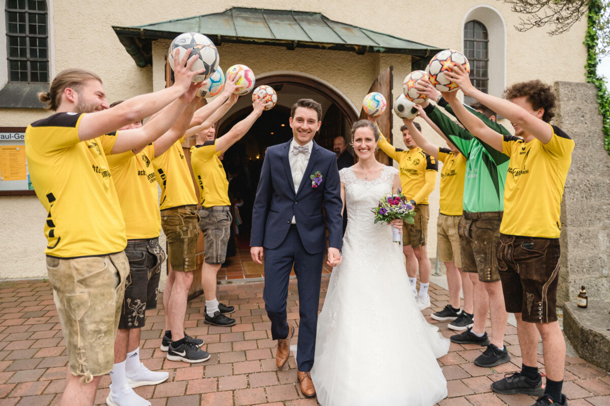 Ein Paar verlässt ein Gebäude und wird von einer Gruppe von Menschen in gelben Trikots und Lederhosen begrüßt, die Fußbälle über dem Kopf halten. Die Braut hält einen Blumenstrauß und beide lächeln.