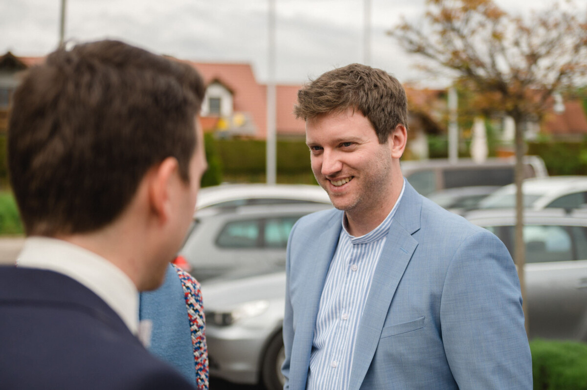 Zwei Männer unterhalten sich im Freien in der Nähe geparkter Autos. Der Mann, der in die Kamera blickt, lächelt und trägt einen hellblauen Anzug.