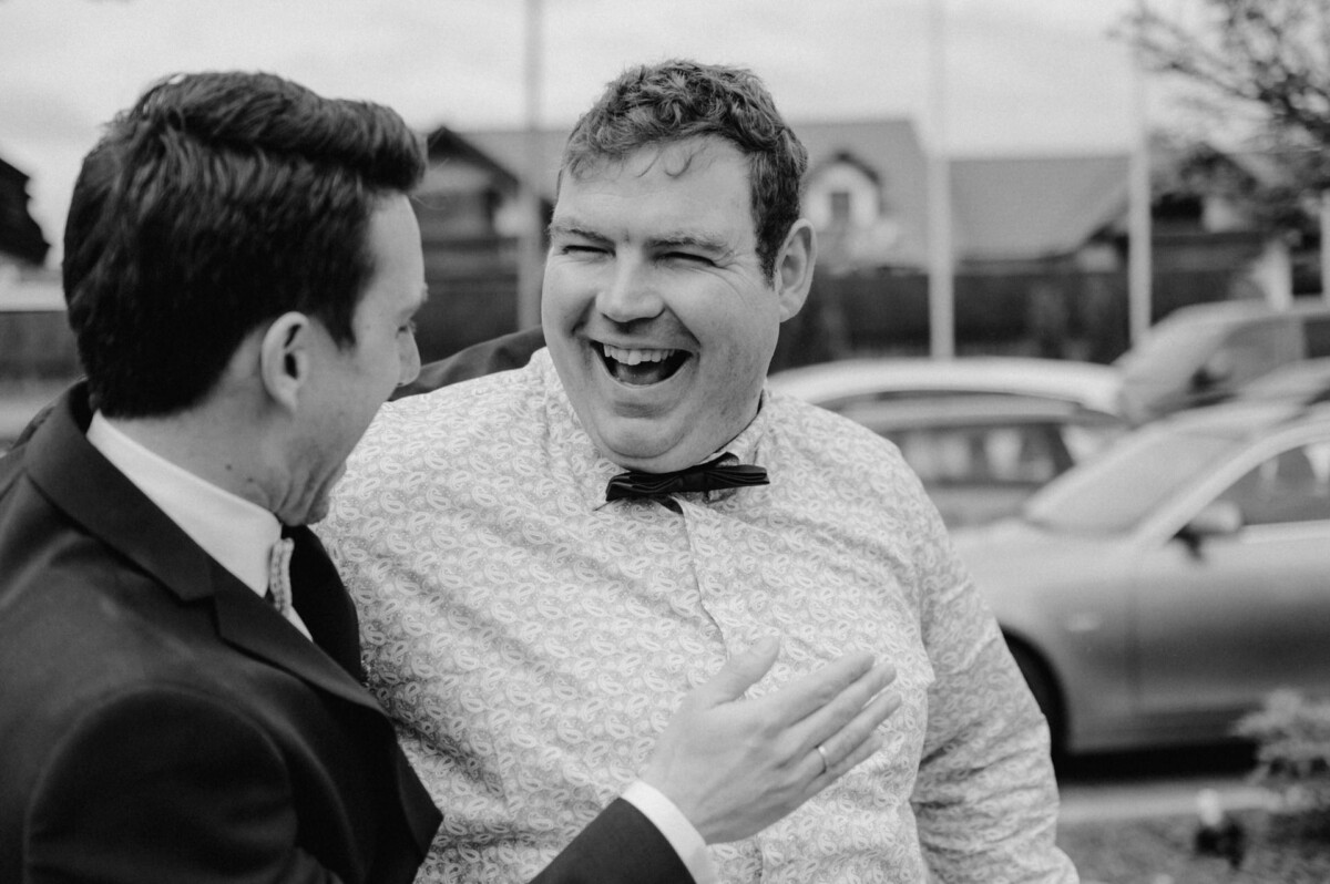 Zwei Männer lachen draußen zusammen auf einem Schwarzweißfoto. Einer trägt Fliege und gemustertes Hemd, der andere einen Anzug. Im Hintergrund sind Autos und Häuser zu sehen.