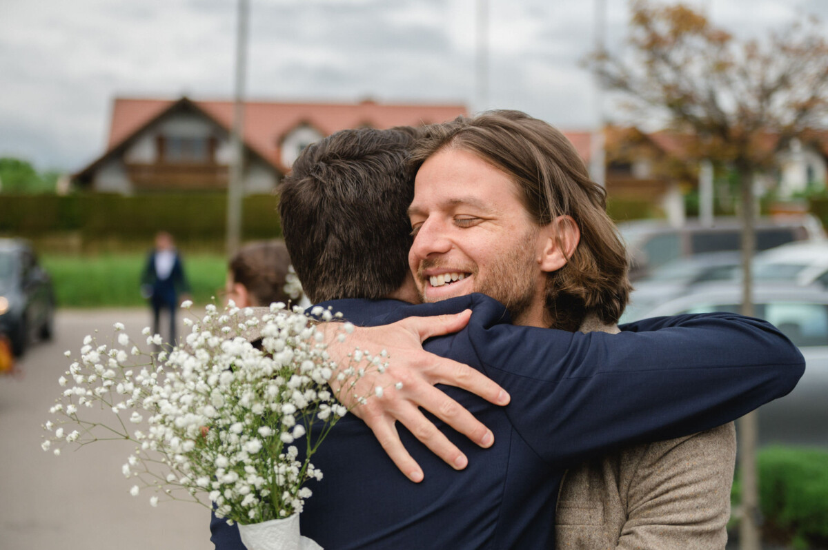 Zwei Menschen umarmen sich im Freien, wobei eine Person einen kleinen Strauß weißer Blumen hält.