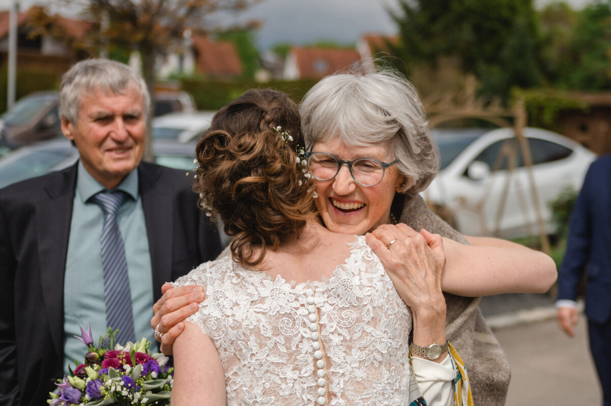 Eine ältere Frau mit Brille umarmt eine Braut, die ein Spitzenkleid trägt. Ein Mann im Anzug steht daneben. Sie sind draußen, im Hintergrund sind Autos und Bäume zu sehen.