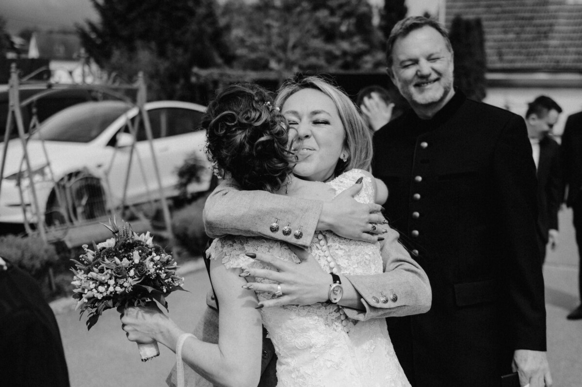 Eine Braut umarmt einen Gast, der einen Blumenstrauß hält, während ein lächelnder Mann in der Nähe steht.