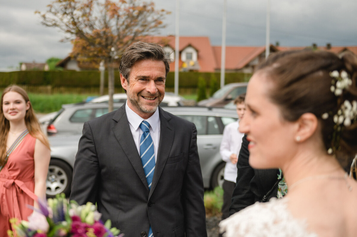 Ein Mann im Anzug lächelt eine Person im weißen Kleid an, im Hintergrund sind andere Menschen und geparkte Autos zu sehen.