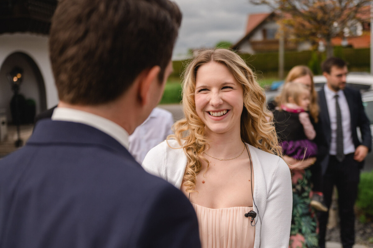 Eine Frau mit lockigem Haar lächelt einen Mann im Anzug im Freien an, umgeben von Menschen in Abendgarderobe.