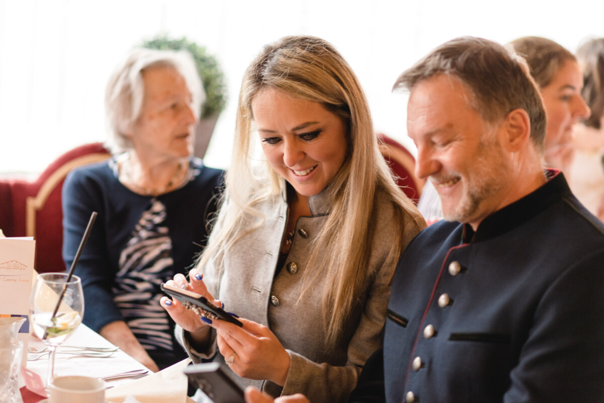 Eine Gruppe von Menschen sitzt an einem Tisch in einem Restaurant. Eine Frau und ein Mann schauen auf ein Gerät, während eine ältere Frau daneben sitzt. Auf dem Tisch sind Weinglas und Speisekarte zu sehen.