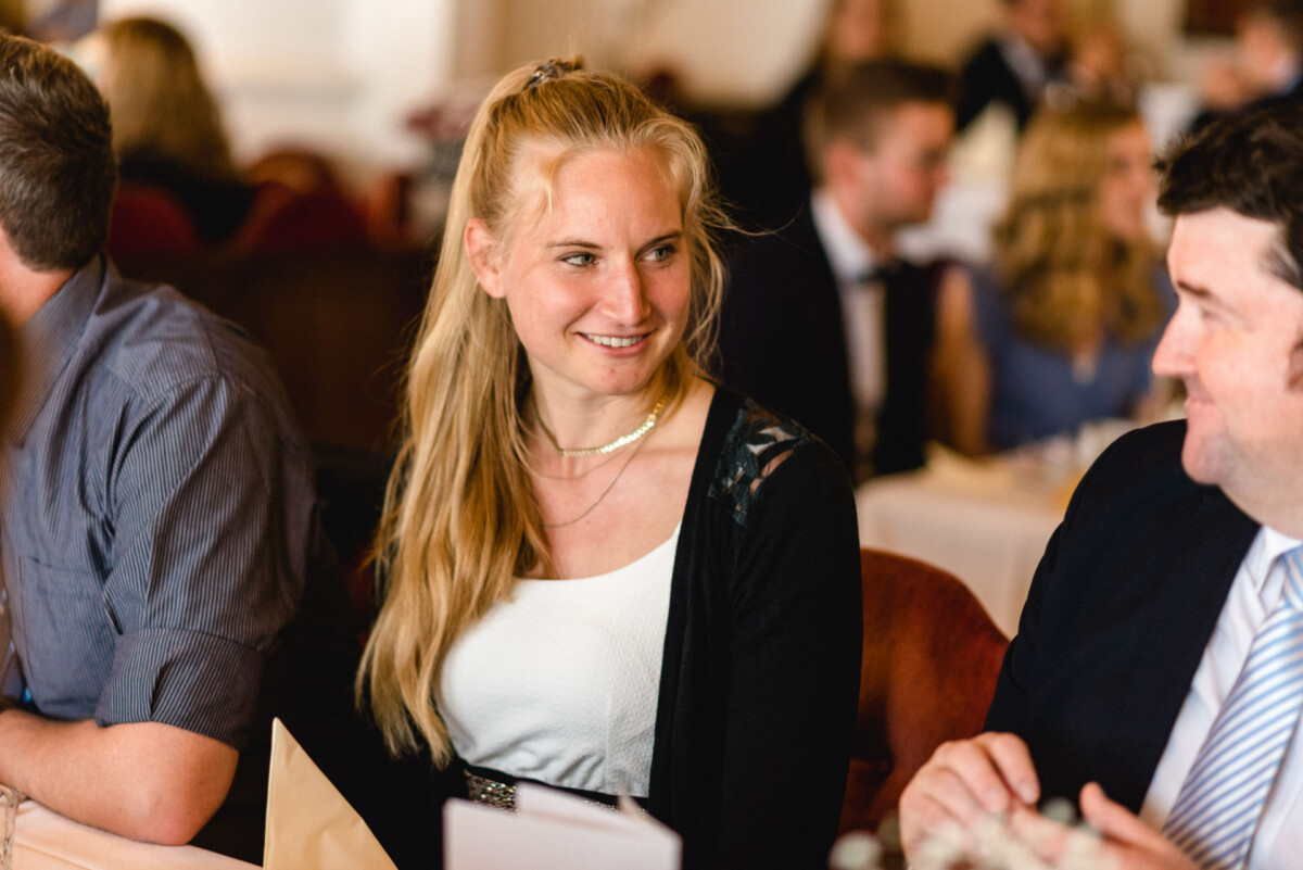 Eine Frau mit blondem Haar sitzt lächelnd an einem Tisch und trägt eine schwarze Strickjacke über einem weißen Oberteil. Um sie herum sitzen Menschen vor einem unscharfen Hintergrund.