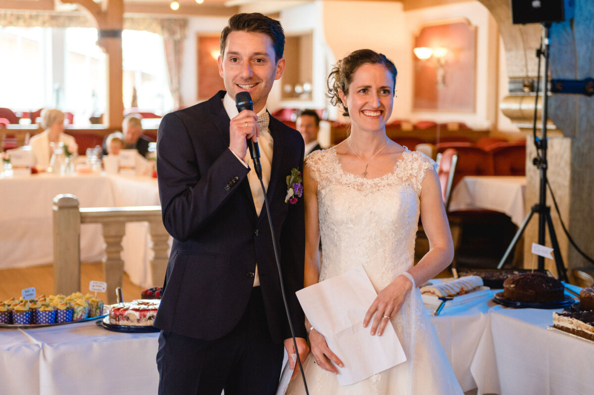 Ein Paar in formeller Kleidung steht zusammen bei einer Hochzeitsfeier. Der Mann spricht in ein Mikrofon und die Frau hält ein Stück Papier. Im Hintergrund ist ein Desserttisch zu sehen.