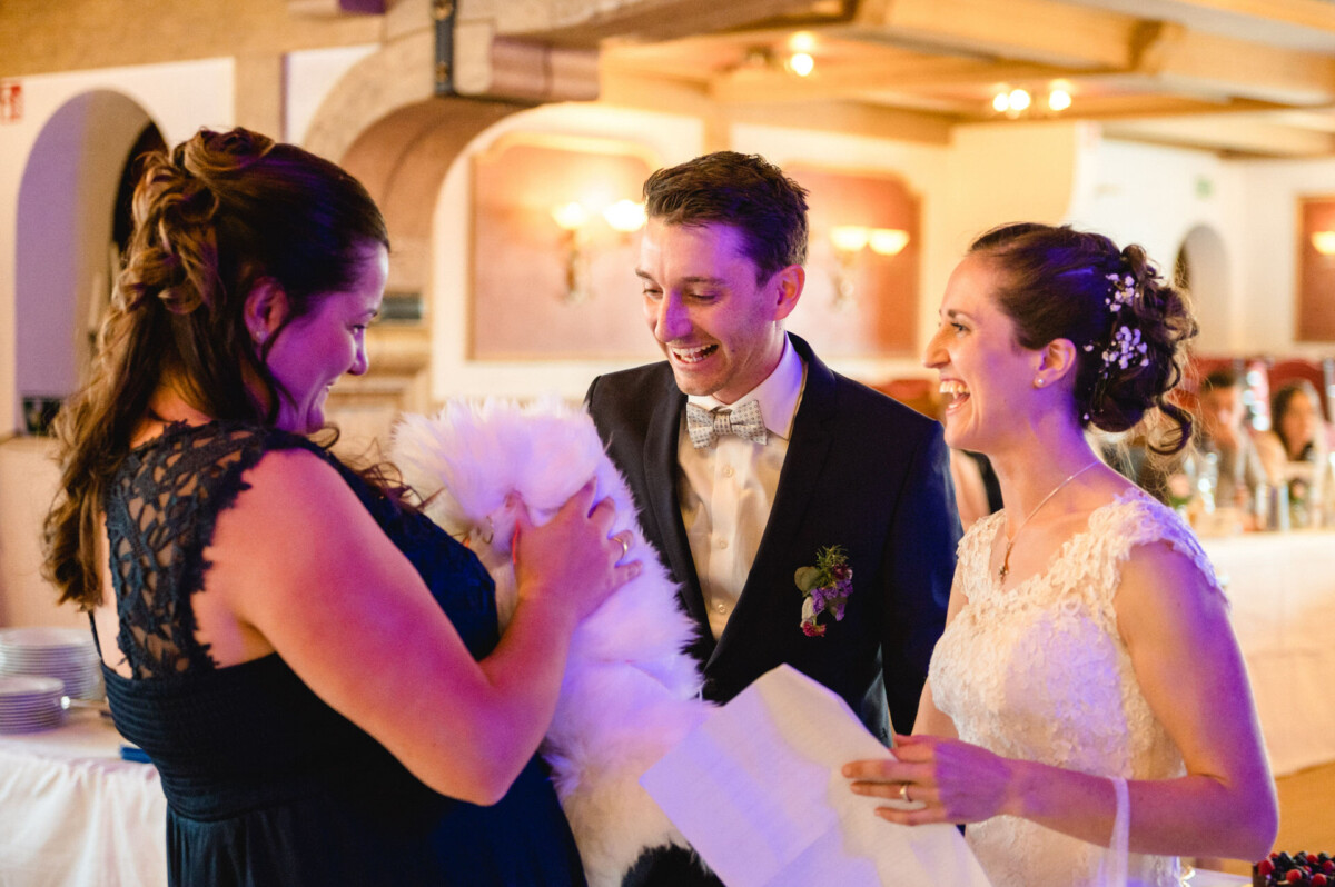 Braut und Bräutigam lächeln, während sie in einem warm beleuchteten Raum mit einem Gast interagieren, der einen flauschigen Gegenstand hält.