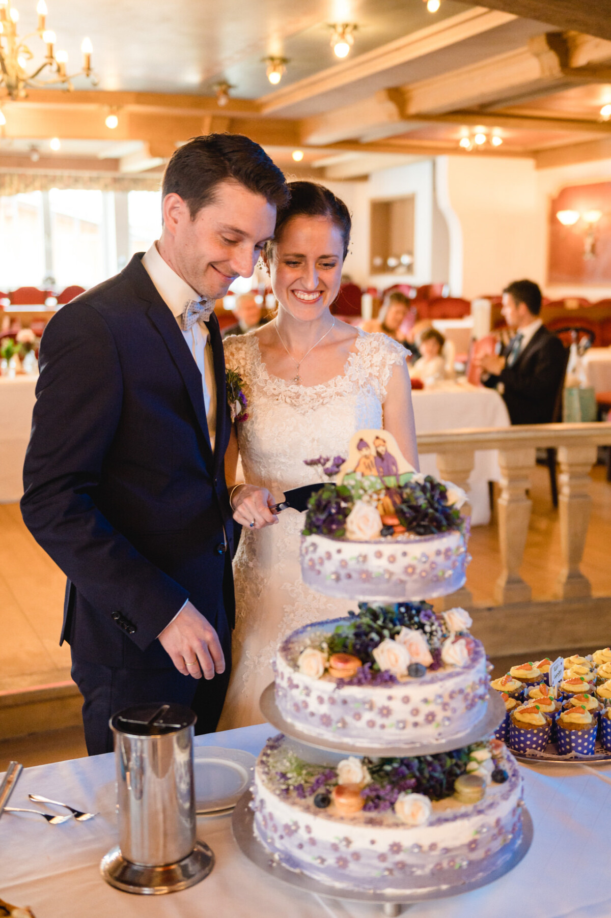 Ein Paar im Hochzeitskleid steht neben einer dekorierten dreistöckigen Torte in einem Empfangssaal. Sie lächeln, im Hintergrund sind Gäste zu sehen.