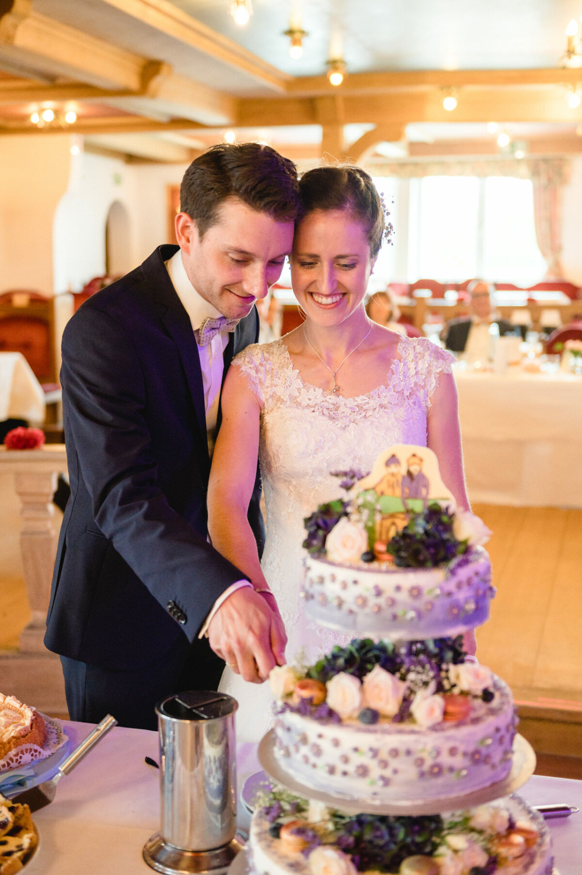 Braut und Bräutigam schneiden in einem Bankettsaal eine mehrstöckige Hochzeitstorte an, die mit Blumen und einer Figur als Aufsatz dekoriert ist.