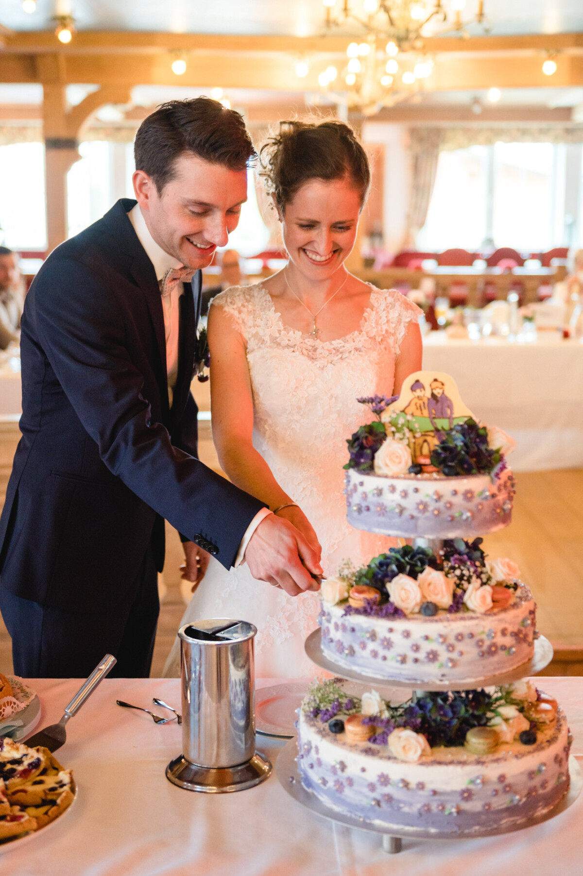 Ein Brautpaar schneidet eine dreistöckige Hochzeitstorte an, die mit violetten und weißen Blumen und einem Brautpaar-Tortenaufsatz dekoriert ist.