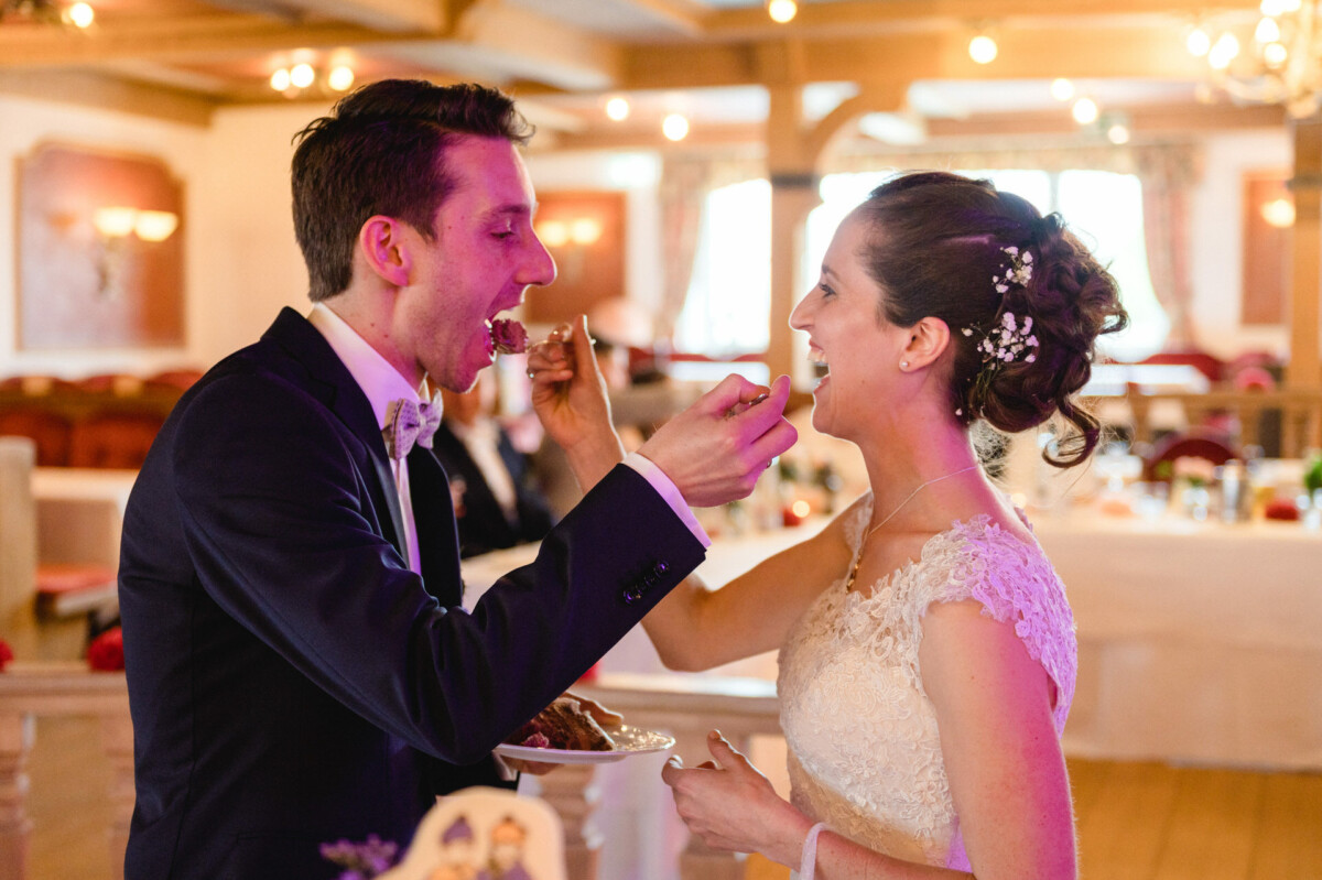 Braut und Bräutigam füttern sich gegenseitig mit Kuchen in einem warm beleuchteten Raum, umgeben von Holzdekor und leeren Stühlen.