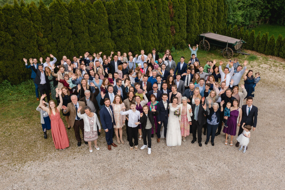 Eine große Gruppe von Menschen, darunter ein Paar im Hochzeitskleid, posiert im Freien mit fröhlichem Gesichtsausdruck und erhobenen Händen.