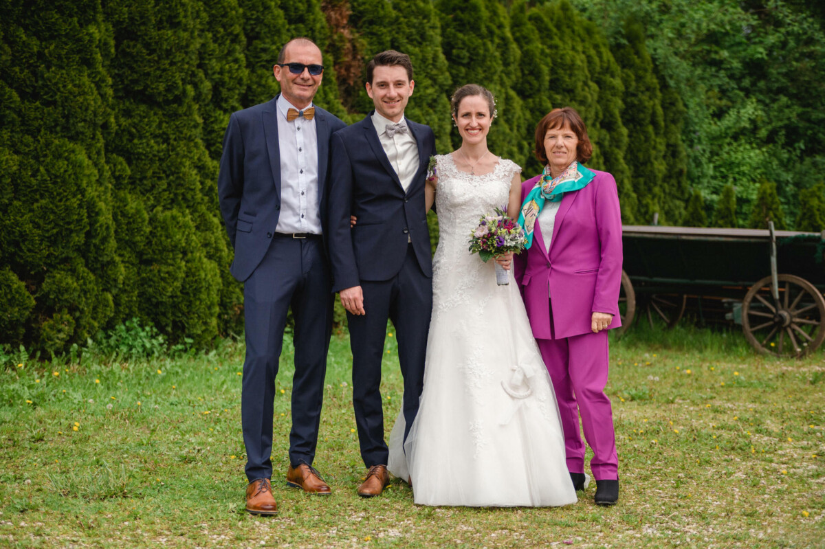 Vier Personen posieren für ein Foto im Freien: zwei Männer in Anzügen, eine Frau in einem Hochzeitskleid mit Blumen in der Hand und eine weitere Frau in einem lila Outfit. Im Hintergrund sind Bäume und ein Karren zu sehen.