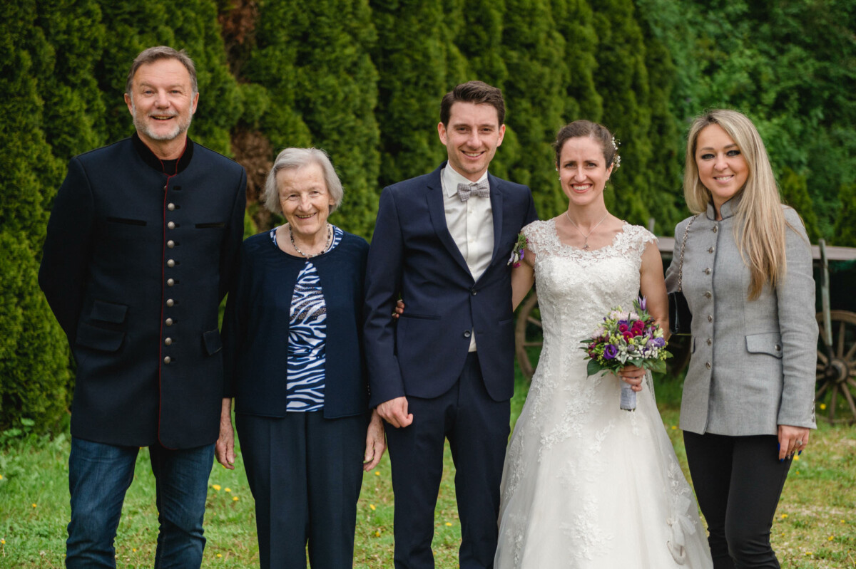 Eine Gruppe von fünf Personen steht im Freien, darunter eine Braut in einem weißen Kleid mit Blumen in der Hand, ein Bräutigam im Anzug und drei weitere Personen in legerer Abendgarderobe. Im Hintergrund sind Bäume und Gras zu sehen.